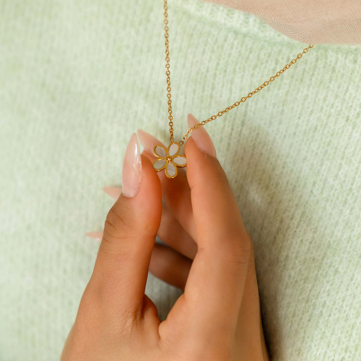 Hand holding a gold necklace with a floral pendant against a light green background