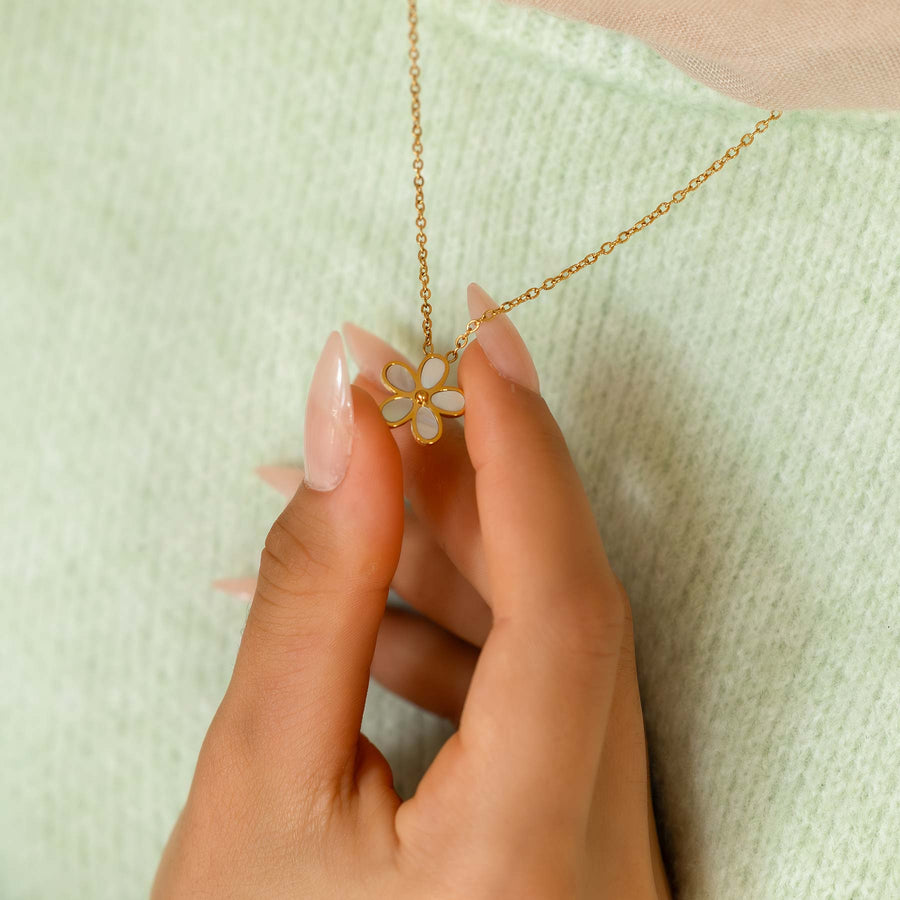 Hand holding a gold necklace with a floral pendant against a light green background